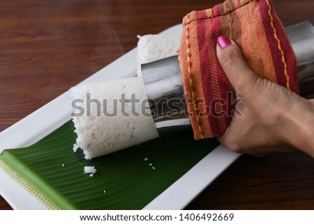 Woman pushing hot Puttu or Pittu holding metal Puttu Kutti vessel in hand. Popular South Indian steamed breakfast dish made of rice flour, grated coconut Kerala, India. Sri lankan food on banana leaf.