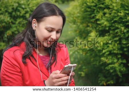girl in a red jacket in the Park listening to music with headphones earbuds with a smartphone in hand.