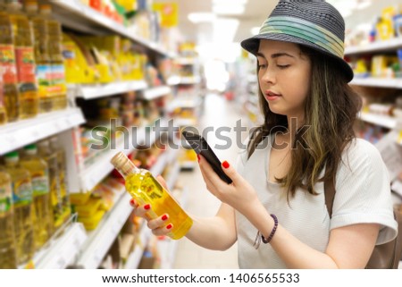A young beautiful woman holds a bottle of oil in her hand and takes a picture of her on a mobile phone. In the background shelves with products. The concept of modern shopping in the store.