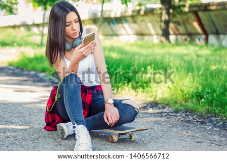 Girl sitting on skateboard and use mobile phone on street. Outdoors, urban lifestyle.
