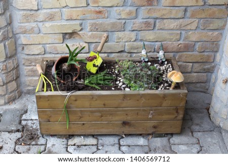 Wooden flower pot with plants and gardening tools in front of brick wall