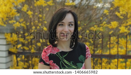 Attractive young woman in a dress with flowers making funny faces near fence. Cute girl with black hair. Close-up shot