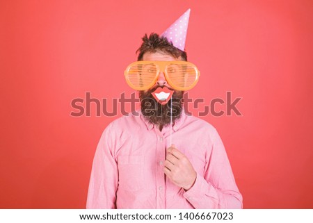 Guy in party hat celebrates, posing with photo props. Hipster in giant sunglasses celebrating. Man with beard on cheerful face holds smiling mouth on stick, red background. Photo booth fun concept.