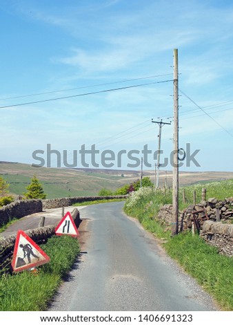 men at work and lane narrowing signs on a narrow country lane with wooden electricity poles and lines in a sunlit rural landscape on the old howarth road in calderdale west yorkshire