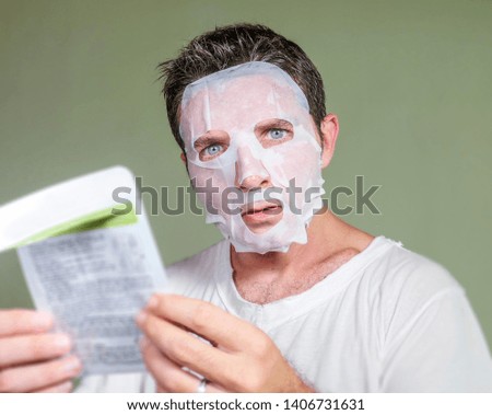 lifestyle isolated portrait of young weird and funny man at home trying using beauty paper facial mask cleansing learning anti aging facial treatment reading the product instructions 