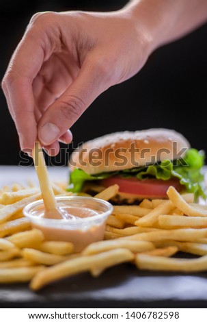 gourmet hamburger with french fries and dipping sauce on a slate cutting board and white red checkered paper, caucasian woman hand dipping french fry 