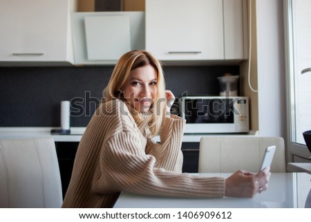 beautiful young woman in a cozy sweater is sitting in the kitchen using a video call in the messenger. Girl at home with a phone in her hands listening to music and talking on the phone with friends