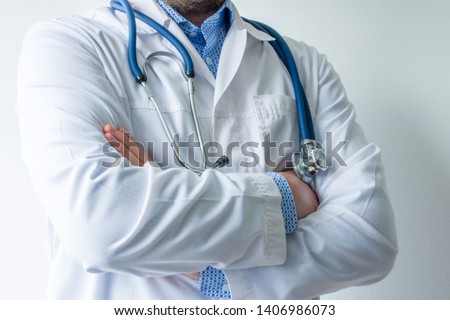 Photo of body of doctor in white lab coat with stethoscope on his neck in half-turn with folded hands against white wall in hospital with light source on left. Concept image of  medical worker 