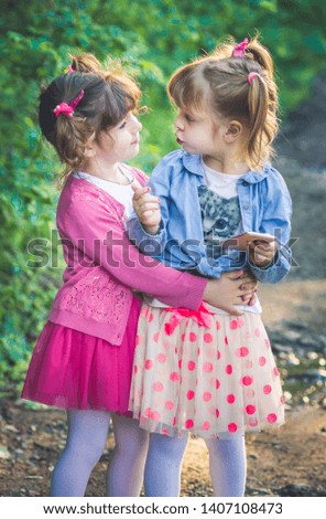 Two little girls, sisters in pink dresses with piggy tail playing with smart phone, looking at each other. Nature, outdoors. Modern child activity concept