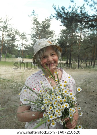 A pretty blond woman in a white dress with a pink pattern and a hat is standing in the forest with a bouquet of daisies in the hands