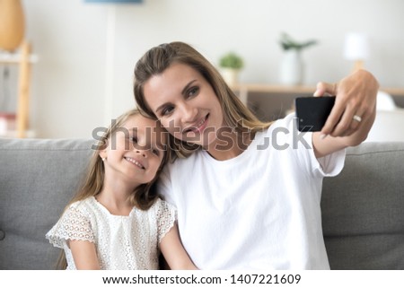 Attractive female sitting together on couch with little preschool girl spend free time in living room at home. Positive mother and daughter using gadget mobile phone smiling taking selfie photography