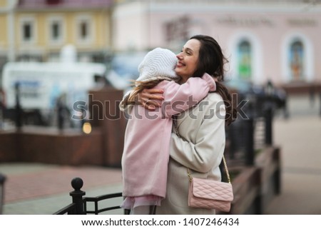 Daughter and mother with long hair in a coat hug on the street of the city, mother kisses and hugs a girl 5 years old, spring city