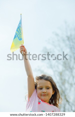 Ukraine s blue-yellow flag flying in wind in hands of little Ukrainian girl on Day of ndependence of Ukraine. Symbols of Ukraine in hands of a smiling child against blue sky and yellow grass.
