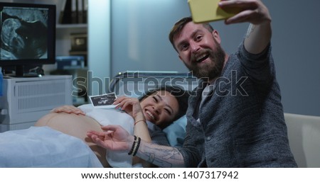 Medium shot of a happy couple taking a selfie with ultrasound photo of their future child in sonography cabinet