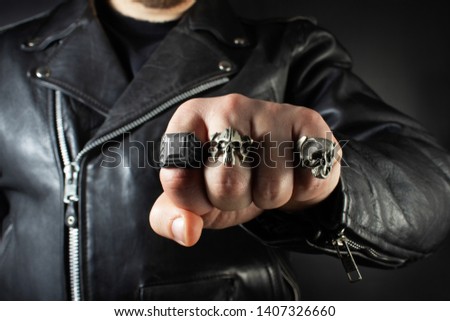 Photo of a man in black leather biker jacket showing fist with skull rings closeup view on black background.