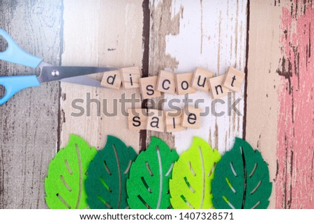 Concept image of price cut. Discount sale text being cut by blue color scissor over nice wooden texture background and green leaves prop