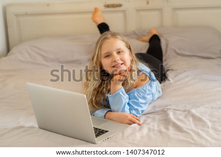 Beautiful cute happy blonde girl playing and surfing the internet on laptop. Child watching cartoon or video on laptop on the bed at home. Digital technology education and children internet usage.
