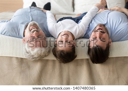 Portrait of happy three generations of men lying on bed upside down looking at camera smiling, little son, father and grandfather have fun enjoy leisure time together posing for picture at home