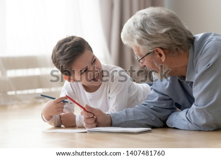 Smiling little boy lying on floor having fun enjoy painting in album together with loving grandfather, grandparent spend time teaching drawing picture entertaining at home with excited small grandson