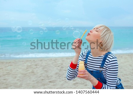 portrait of beautiful young pretty, happy, cute, blonde woman in striped shirt, denim overalls with soap bubbles by the sea