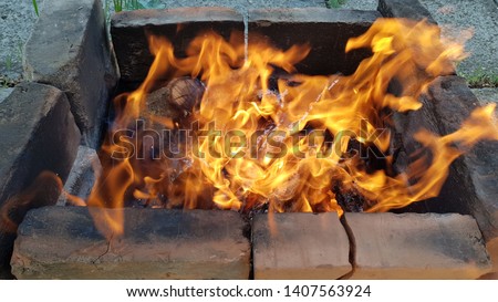 Bonfire with bright orange flame in old firewood brazier made from burnt smoky bricks into ground. Fire is flaming in windy weather. Blurry blazing campfire structure closeup.