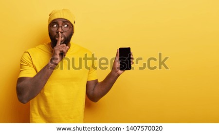 Embarrassed black man makes shush gesture, tries be speechless, looks secretly aside, holds modern cell phone device with mock up screen, tells secret functions of cellular, isolated on yellow wall