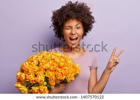 Funny young woman blinks eyes, makes peace sign with hand, expresses happiness, holds orange tulips, wears casual violet t shirt poses indoor. Afro American celebrates birthday isolated on purple wall