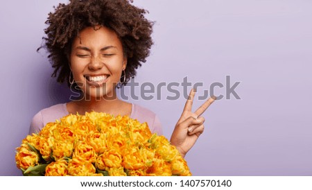Headshot of optimistic Afro woman feels energized and amused, demonstrates victory gesture, holds orange tulips, keeps eyes shut, has broad shining smile, isolated over purple background, copy space