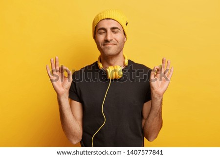 Headshot of satisfied male model makes okay gesture with both hands, has eyes closed, meditates indoor alone, wears casual black t shirt, wears headphones on neck, isolated on yellow studio wall
