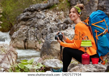 Young backpacker or hiker has rest on stone after walking long distance, enjoys journey through undisturbed nature, makes coffee on special tourist equipment, uses camera for taking pictures