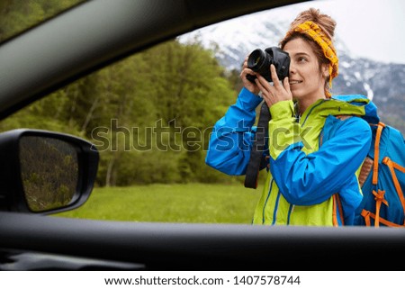 View from car of professional young woman photographer takes pictures on camera, walks on green field with mountain landscape, has country hike, makes travel nature photography, wears jacket