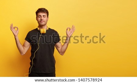 Concentrated young European man keeps hands in okay gesture, meditates indoor, has eyes closed, tries to relax with calm music in headphones, dressed in casual outfit, isolated on yellow wall