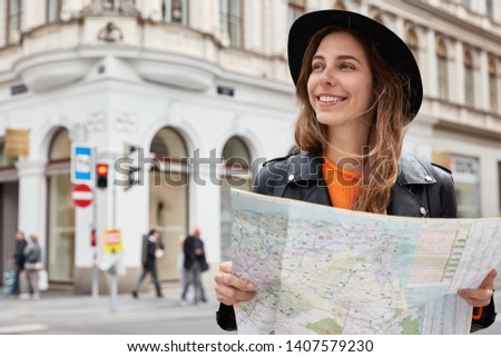 Portrait of female traveler stands with paper map, searches for attractions in big famous city, looks aside with smile, wears black headgear, strolls around new place, admires beautiful sights