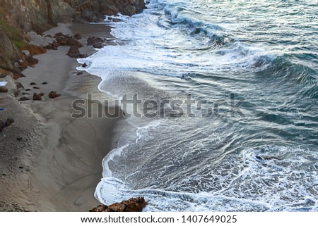 breaking waves and foam trails on a dark sandy beach with rocks and cliff edge