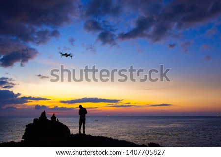 Silhouette of young man using and watching a flying drone in twilight sky over the sea for photos or video making.