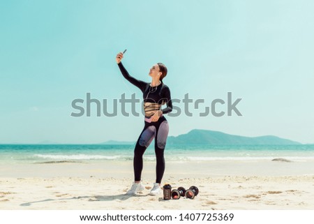Cheerful young sportswoman with headphones taking a selfie with outstretched hands while standing at the beach. Phuket. Thailand. Summer vacation and sport activity