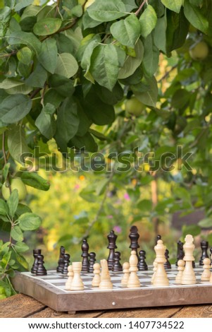 Chess board with chess pieces on wooden desk with branches of apple tree and green leaves on the background. Selective focus on white pieces.