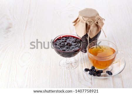 Honeysuckles berries, jam in a bowl, cup of tea and glass jar of jam stand on a wooden background.