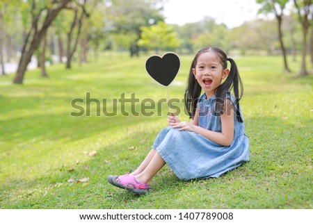 Happy little Asian kid girl holding blank heart label sitting on green grass at garden outdoor.