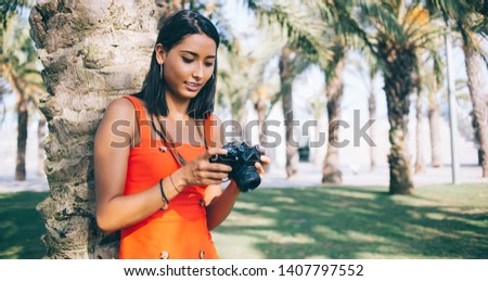 Young hipster girl holding retro equipment in hands and recalls to pleasant moments while rewatching photos, Latin millennial amateur photographer with vintage camera standing near palm tree