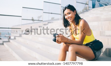 Portrait of excited female photographer sitting on city stairs with favourite old fashioned equipment in hands and smiling at camera, cheerful rejoice Latin woman amateur with retro technology