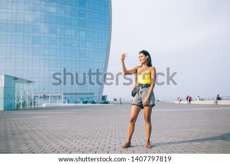 Positive hipster girl posing and clicking selfie photo on smartphone front camera enjoying summer trip for discover world, happy millennial female tourist taking pictures via mobile phone