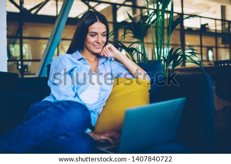 Positive smiling hipster girl searching funny movie for watching using modern laptop device indoors, happy woman resting on comfortable sofa while messaging with friends via application on computer