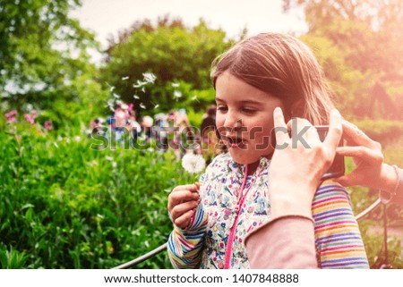 Cheerful mother taking picture of her cute little daughter in the park blowing dandelion