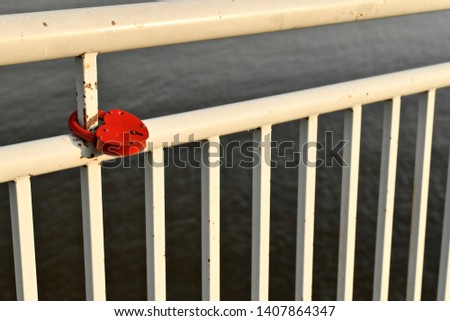 White painted seafront railing. With a red lock in the shape of a heart, on a metal pipe. Diagonal view of the lattice against the background of the river. Sunset light,