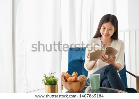 Young beautiful Asian woman relaxing in living room at home, reading books, drinking coffee in the morning with happy feeling