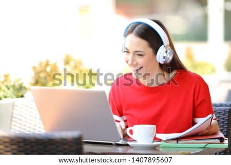Happy student with headphones e-learning with a laptop sitting in a coffee shop terrace