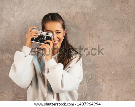 Beautiful young smiling photographer girl taking photos using her retro camera. Woman making pictures. Model dressed in casual summer hoodie clothes. Posing In studio near gray wall