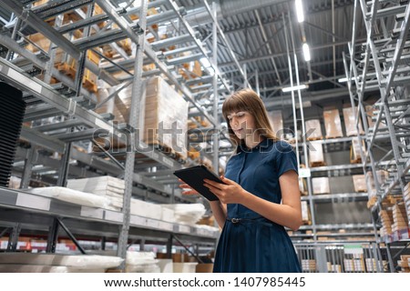 Wholesale warehouse. Beautiful young woman worker of store in shopping center. Girl looking for goods with a tablet is checking inventory levels in a warehouse. Logistics concept Royalty-Free Stock Photo #1407985445
