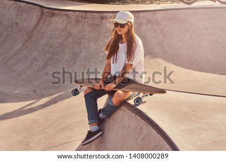 Beautiful young woman in sunglacces and cap is sitting at skatepark on the ramp with her longboard. 